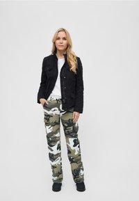 Giacca di suede nera sopra una maglietta bianca, abbinata a pantaloni cargo mimetici verde e bianchi e stivaletti neri. Outfit testurizzato e informale.