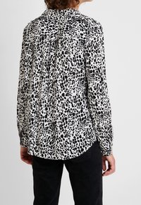 Blusa a maniche lunghe in tessuto leggero, caratterizzata da un motivo a pois bianco e nero, schiena plissettata e polsini con bottoni. Orlo arrotondato.