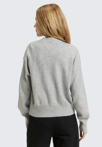 Pull en maille grise avec un ourlet et des poignets côtelés, présentant une coupe décontractée et une surface légèrement texturée. Porté avec un pantalon noir.