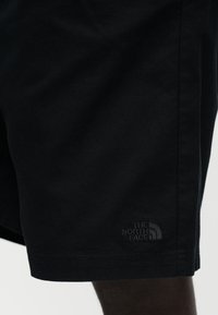 Κοντινή λήψη μαύρου σορτς με το λογότυπο "The North Face" κοντά στο τελείωμα, σε σκουρόχρωμο πόδι με φωτεινό φόντο.