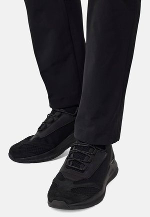 Chaussures de sport noires avec des matériaux en mesh et en suède, présentant un design texturé et un système de laçage, accompagnées de pantalons noirs cintrés.