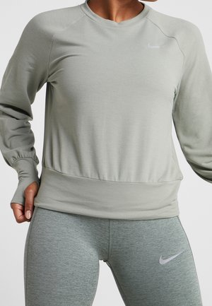 Personne portant un sweat-shirt Nike à manches longues gris clair et un legging Nike gris assorti, main reposant sur la hanche, sur un fond uni.