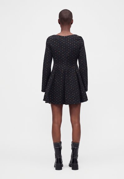 Personne aux cheveux courts portant une robe noire à manches longues avec un petit imprimé floral coloré et des bottines noires, debout dos à la caméra.