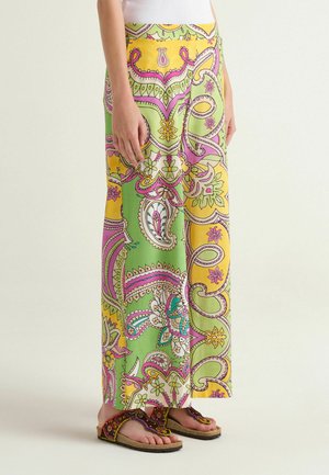 Frau trägt weit geschnittene Hosen mit buntem Paisley-Muster in Grün, Gelb, Pink und Weiß, kombiniert mit Sandalen mit Perlen.