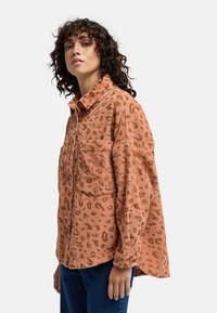 Camicia in velluto a coste marrone con stampa leopardata, chiusura frontale con bottoni, due tasche sul petto e vestibilità rilassata. Presenta un colletto e maniche con polsini.