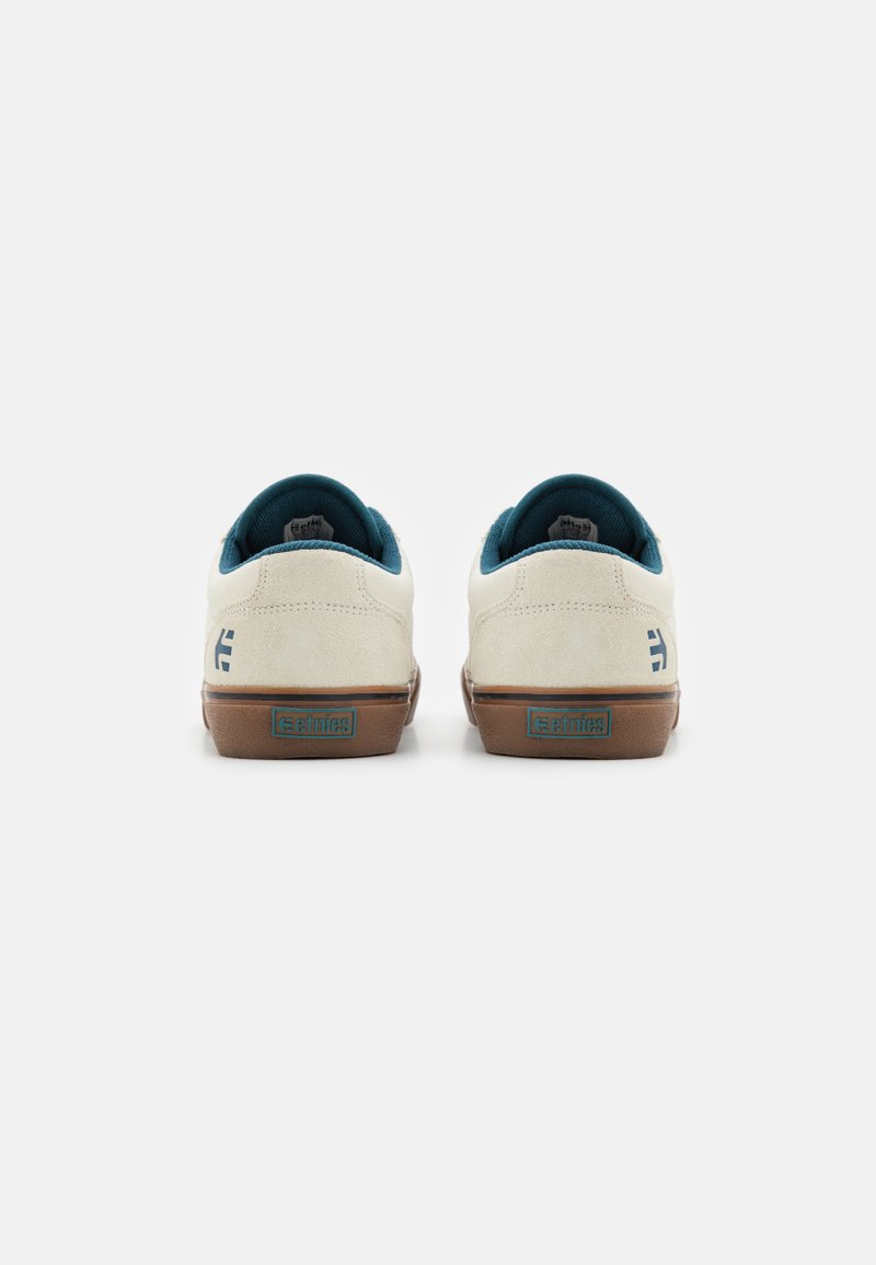 Weiße Wildledersneaker mit einer braunen Gummisohle, türkisem Innenfutter und einem kleinen blauen Logo an der Ferse, das ein sauberes, minimalistisch gestaltetes Design aufweist.