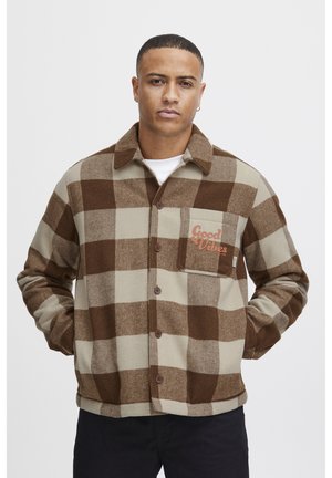 Pruun ja beež ruuduline flanellshirt, millel on nööbist sulgemine ja rinnaõmmeldud tasku, kus on oranž tekst "Head vibratsioonid".
