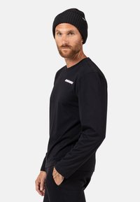 Schwarzes Langarmshirt mit einem Rundhalsausschnitt und einem pinken Logo auf der linken Brust. Das Model trägt eine schwarze gerippte Mütze.