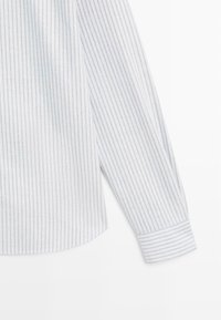 Biała koszula z długim rękawem w wąskie niebieskie paski. Miękka bawełniana tkanina, mankiet zapinany na guziki, zaokrąglony dół i widoczne detale szwów.