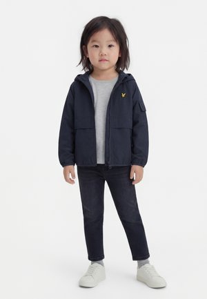 Dziecko stoi przodem, ubrane w granatową kurtkę z kapturem, szarą koszulkę, ciemne dżinsy, szare skarpetki i białe trampki na jednolitym białym tle.