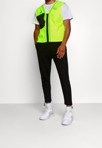 Neongrön och svart zippered väst med axel- och bröstfickor. Svarta byxor med dragkedjor vid fotlederna. Vita sneakers med mönstrade strumpor.