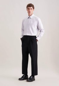 Camicia a maniche lunghe grigio chiaro con tasca, abbinata a pantaloni neri plissettati e scarpe eleganti nere, indossata da un uomo con le mani nelle tasche.