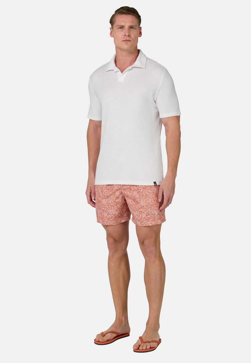 Polo-shirt blanc avec col, associé à un short à motifs orange. Le modèle se tient en tongs, montrant une tenue d'été décontractée.