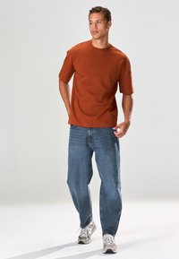 Kortärmad t-shirt i rostfärg kombinerad med lösa blå jeans. Modellen har på sig ljusgrå sneakers med vita skosnören. Enkel design och passform.