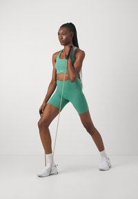 Zeleni športni crop top in kratke hlače z visokim pasom, izdelani iz elastičnega materiala. Model uporablja elastične trakove in nosi bele superge.