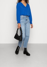 Blouse bleu roi à manches longues avec un col en V, associée à un jean droit bleu clair. Sac noir avec une poignée torsadée et bottines noires épaisses.