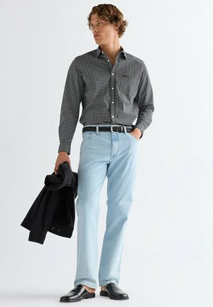 Mężczyzna ubrany w czarno-białą koszulę w kratę, jasnoniebieskie dżinsy, czarny pasek, czarne buty wsuwane, trzymający czarną kurtkę, stojący na białym tle.