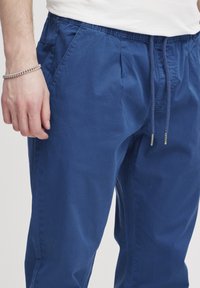 Niebieskie bawełniane spodnie z elastycznym pasem i sznurkiem ściągającym. Posiadają przeszyte zakładki i boczne kieszenie. Gładka tekstura.