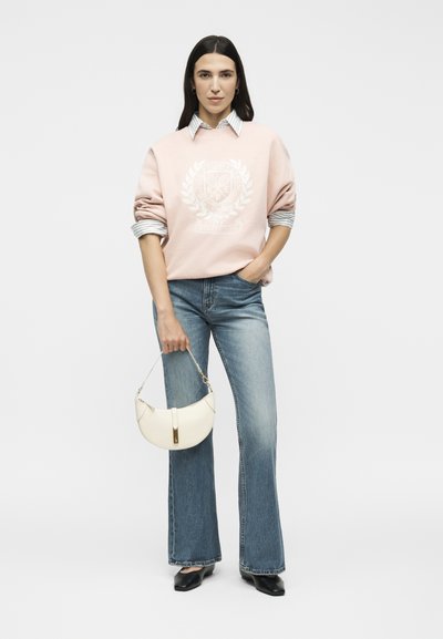 Femme portant un sweat-shirt rose par-dessus une chemise rayée, un jean évasé bleu, des chaussures noires, tenant un sac à main blanc avec un fermoir doré.