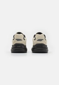 Sneaker mit einer schwarzen Gummisohle, ausgestattet mit einem strukturierten beigen Obermaterial und schwarzen Akzenten, die ein modernes, sportliches Design zeigen.