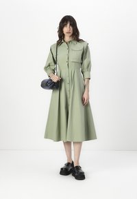 Rochie verde deschis, cu nasturi, cu un corset ajustat și fustă evazată, având un buzunar pe piept și mâneci lungi. Împărată cu o geantă neagră și pantofi cu platformă.