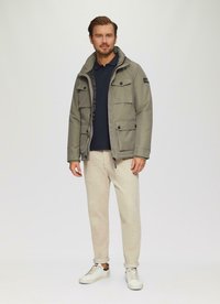 Olivegrüne, kapuzenjacke mit mehreren Fronttaschen und einem Reißverschluss. Kombiniert mit hellbeigen Hosen und weißen Sneakern, die graue Akzente aufweisen.
