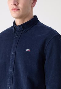 Camicia in corduroy blu navy con colletto button-down, caratterizzata da un piccolo logo ricamato rosso e bianco sul lato sinistro del petto. Tessuto testurizzato.