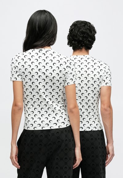 T-shirts brancas de manga curta com um padrão de lua crescente preta, usadas por duas pessoas, combinadas com calças escuras com padrão.