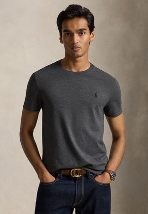 Jeune homme aux cheveux noirs courts portant un t-shirt gris foncé, un jean bleu, une ceinture marron et une montre-bracelet, debout devant un fond uni.