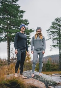 Långärmade termotröjor och figurnära leggings i mörkblått och grått, tillverkade av slätt tyg, barfota på stenig terräng omgiven av träd.