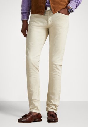 Mand iført cremefarvede bukser, brune læder tassel loafers, lilla sweater, brun vest og armbåndsur med den ene hånd i lommen.