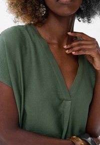 Oliwkowa, krótko rękawowa bluzka z dekoltem w serek. Miękka tekstura, luźny krój, z centralnym szwem. Uzupełniona wzorzystymi kolczykami i bransoletkami.