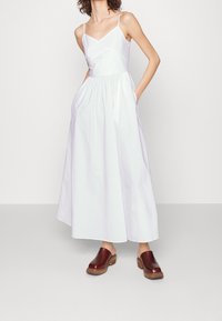Weißes Baumwollkleid ohne Ärmeln mit V-Ausschnitt, gerafftem Taillenbund und seitlichen Taschen, kombiniert mit braunen Holzclogs.