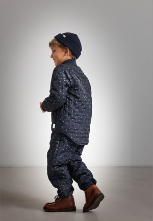 Enfant portant un chapeau bleu marine, une veste à motifs et un pantalon, marchant à l'intérieur avec des bottes en cuir marron sur un sol et un mur gris.