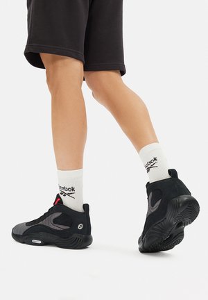 Czarno-szare buty sportowe z teksturowaną cholewką, wyściełanym kołnierzem i gumową podeszwą. Noszone z białymi skarpetkami z logo Reebok.