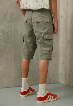Shorts cargo vert olive avec deux poches latérales, des poches arrière et un logo écusson. Portés avec des baskets en daim rouges et des chaussettes blanches.