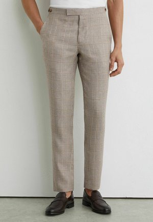 Muž nosiaci béžové a hnede kárované úzke nohavice s prednými záhybmi, v kombinácii s tmavohnedými koženými mokasínmi, stojaci pri bielej stene.