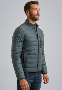 Giacca imbottita con zip di colore verde scuro, caratterizzata da cuciture orizzontali, tasca nera a contrasto e colletto alto. Indossata sopra un paio di jeans in denim.