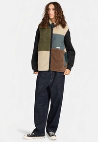 Fleece-Weste mit Patchwork-Design in Grün, Beige, Braun und Blau. Mit Frontreißverschluss, Taschen und schwarzen Ärmeln. Kombiniert mit dunklen Jeans.