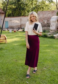 Model wearing a white short-sleeve top and a burgundy skirt, holding a black clutch, standing on green grass with light natural surroundings.