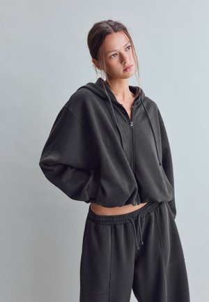 Ung kvinde med håret bundet tilbage iført en sort hættetrøje med lynlås og matchende joggingbukser, der kigger let til siden mod en ensfarvet baggrund.