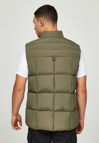 Olivgrön pufferväst med hög krage, quiltad design och en ögla på baksidan. Modellen har en vit t-shirt under.