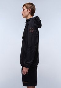 Zwarte lichtgewicht jas met een capuchon, rits aan de voorkant, elastische manchetten en een cirkelvormige logo patch op de linker schouder. Draag over zwarte shorts.