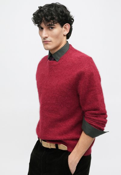 Piros, texturált kötött pulóver, kerek nyakkivágással, sötétzöld, galléros ing felett viselve. A modell keze a zsebében van, bemutatva az öltözéket.