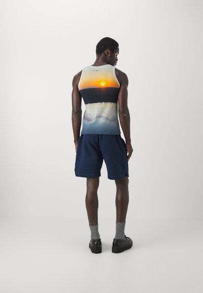 Hihaton tank top, jossa on auringonlaskugrafiikka oransseilla ja tummansinisen väreillä; yhdistetty tummansinisiin shortseihin, harmaisiin sukkiin ja mustiin kenkiin.