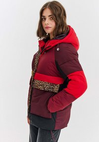 Žena v marónovej a červenej prešívanej bundě s kapucňou a leopardím vzorom, stojaca s jednou rukou v vrecku na jednofarba pozadí.