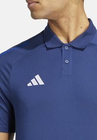 Marineblauwe polo met een gestructureerde afwerking, knoopsluiting en contrasterend wit Adidas-logo op de linkerkant van de borst. Korte mouwen.