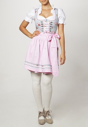 Jeune femme portant une robe traditionnelle dirndl à motifs floraux avec un chemisier blanc, un tablier rose noué à la taille, des collants blancs et des chaussures beiges à lacets.