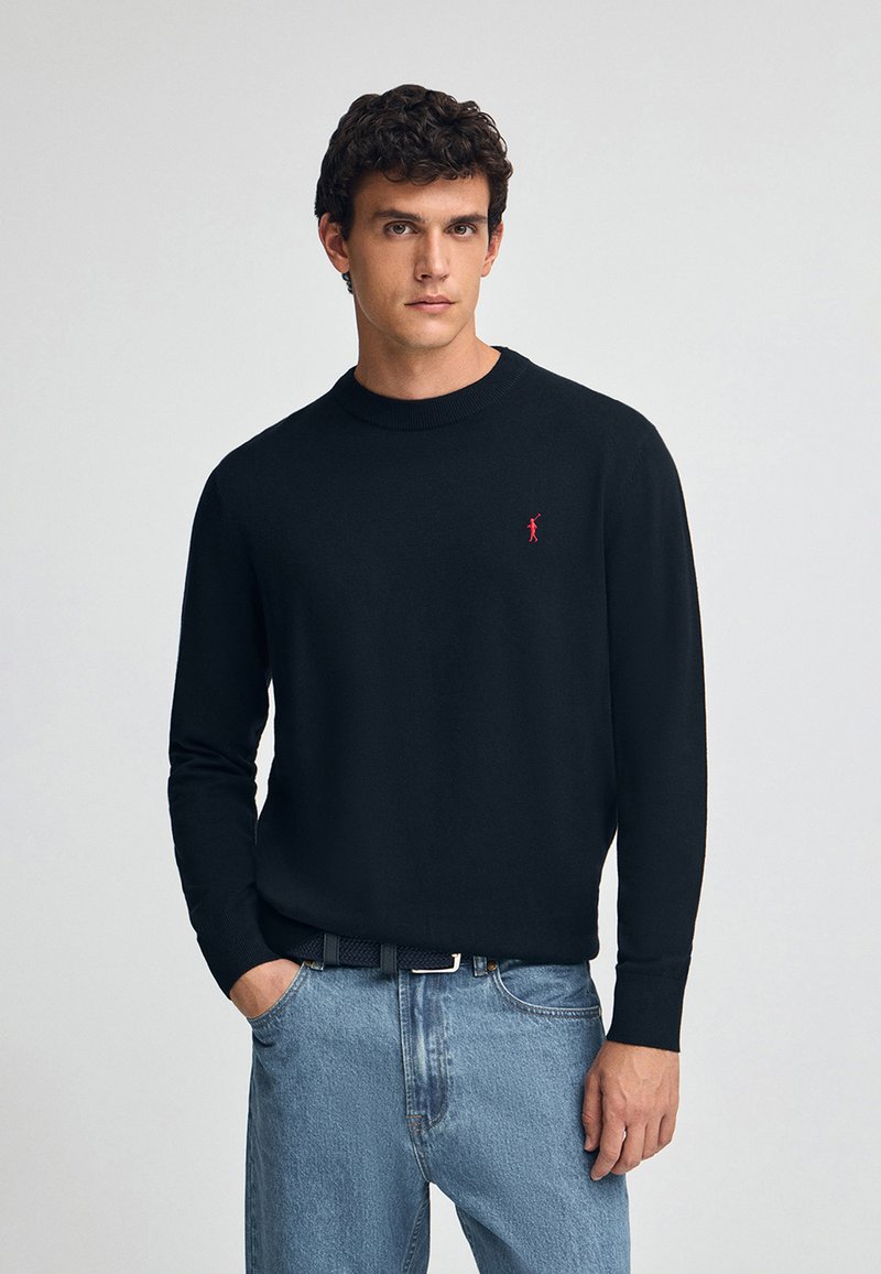Un pull en tricot noir à col rond avec un petit logo rouge sur la poitrine, assorti à un jean en denim bleu clair.