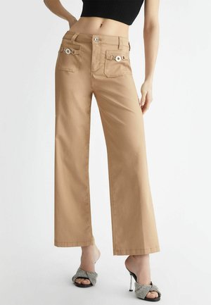 Beżowe spodnie o szerokich nogawkach z przednimi kieszeniami ozdobionymi srebrnymi kółkami, noszone z czarnym krótkim topem i wzorzystymi sandałami na wysokim obcasie.